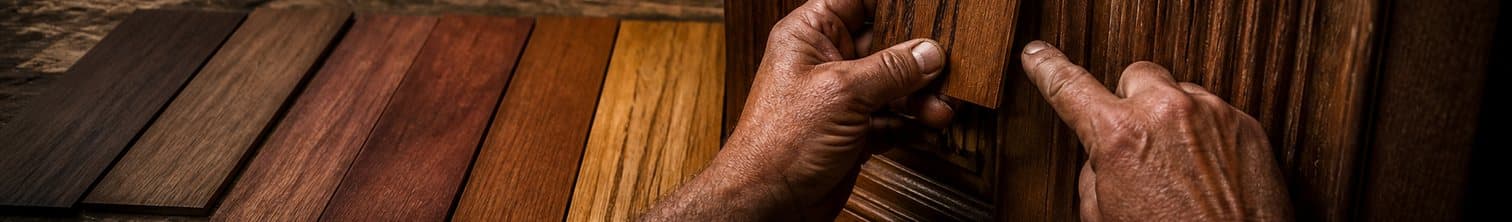 A row of hardwood finish samples fanned across a workbench in a dark workshop, from dark walnut through to honey oak, with one craftsman's hand holding a sample up against the ribbed carving of a door panel to compare the tone and the other pointing at the grain