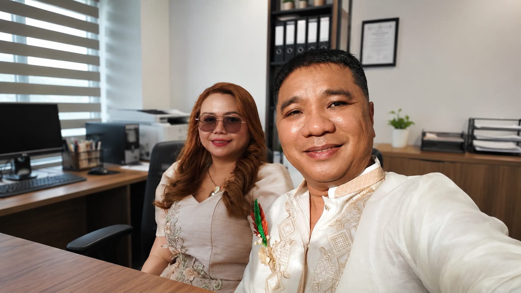 The owners of OLAZO Furniture seated together at the company office, dressed for a formal occasion, with the business's framed certificate on the wall behind them